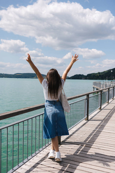 Back view of woman raised her hands up. Female walking through the embankment. Emotion of beauty of nature. Clear water. High quality photo - Photo, Image