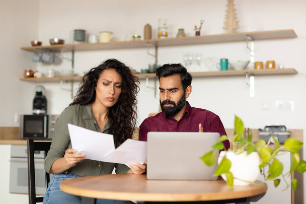 Bankruptcy concept. Overwhelmed arab couple sitting at kitchen desk in front of laptop, holding documents and reading correspondence, got mail from bank - Photo, image