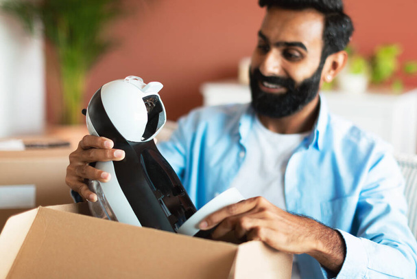 Satisfied Customer. Happy middle eastern man opening cardboard box taking out new gadget at home interior, selective focus on parcel with order. Online shopping and delivery concept - Photo, Image