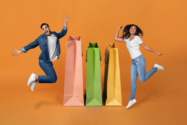 Shopaholisteja. Iloiset Lähi-idän puolisot juhlivat suuria ostoksia, hyppäävät lähellä valtavia laukkuja hymyillen kamerassa Oranssilla taustalla, kollaasi. Ostajat Pari Mainonta Myynti Tarjous Lähellä Shoppers - Valokuva, kuva
