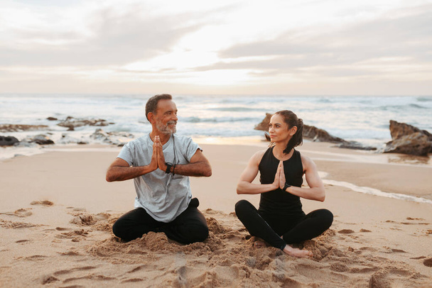 Cheerful senior caucasian lady and man in sportswear practice yoga and breathing, mental exercises, enjoy training and lifestyle on beach in morning, outdoor. Relationships, sports together - Photo, Image