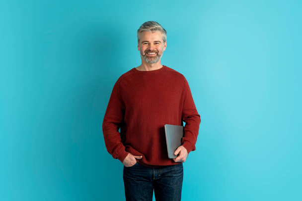Entrepreneurship and technologies. Cheerful handsome middle aged man startup founder or small business owner managing business operations via his laptop, blue studio background, copy space - Foto, afbeelding