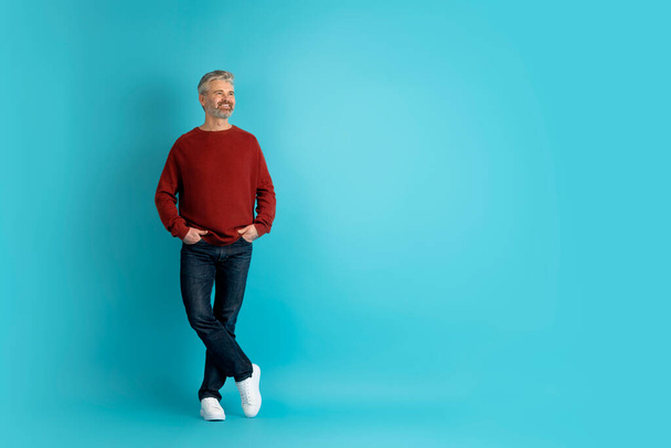 Cheerful positive handsome grey-haired bearded stylish middle aged man wearing casual outfit posing with hands in pockets on blue studio background, smiling and looking at copy space for advert - Photo, Image