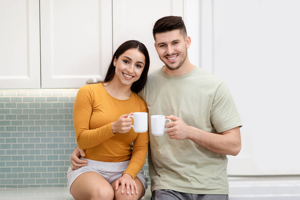 Love, relationships. Portrait of cheerful happy beautiful loving hispanic young man and woman in casual sitting at kitchen, drinking tea or coffee, have conversation, enjoying time together at home - Photo, image