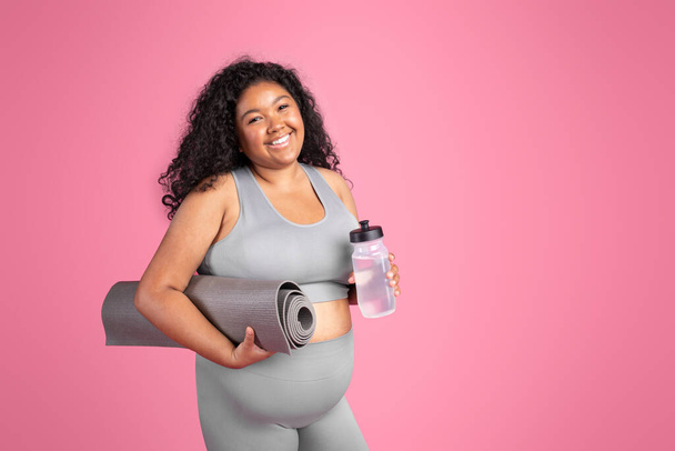 Aufgeregte schwarze Oversize-Lady in Sportbekleidung mit Matte und Wasserflasche, die nach dem Training auf rosa Studiohintergrund ruht, Kopierraum. Empfehlung fitter Lebensstil, Körperpflege - Foto, Bild