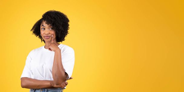 Cheerful pensive millennial black woman in casual looks at blank space, thinking, isolated on orange studio background, panorama. Choice, ad and offer, human emotion, lifestyle - Photo, Image