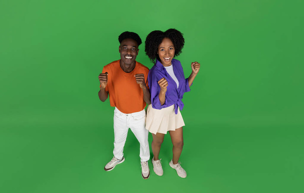 Gülümseyen Afro-Amerikan kadın ve erkek rahat bir şekilde zaferi, başarıyı, yeşil stüdyonun arka planında izole edilmiş manzarayı kutluyorlar. İlişki ve kazanç, satış, sürpriz, iyi haber - Fotoğraf, Görsel