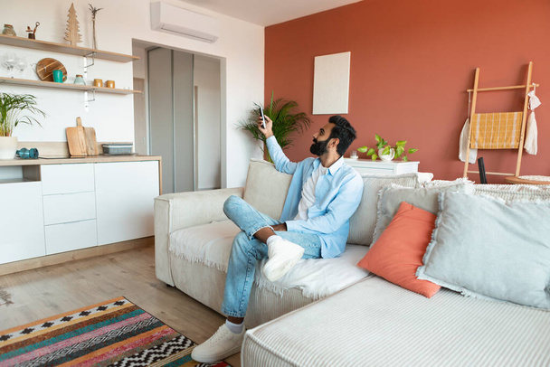 Sistema Dividido. Homem árabe contento segurando controlador remoto relaxante sob o ar condicionado, ajustando o controle do clima e modo de temperatura de ac sentado no sofá na sala de estar interior - Foto, Imagem