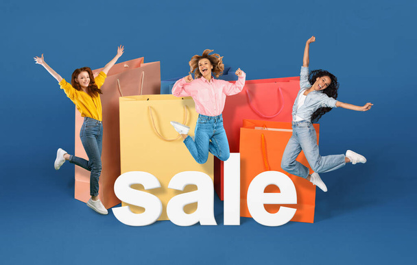 Consumerism. Three women on shopping jumping near large shopper bags and word sale, advertising discount commercial offer over blue studio background. Shopaholics celebating black friday. Collage - Foto, Bild
