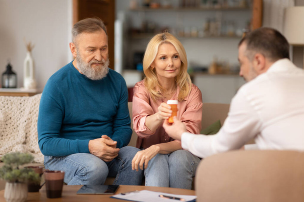 Medical Consultation At Home. Professional Health Advisor Man Giving Pills Bottle To Senior Couple Sitting In Living Room. Spouses Consulting With Specialist About Supplements. Selective Focus - Фото, зображення