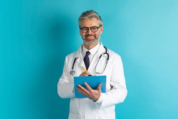 Egészségügyi ellátás, találkozó az orvossal. Boldog ősz hajú, középkorú férfi orvosi fehér kabátban és szemüvegben orvos jegyzetel a jegyzetfüzetben és mosolyog a kamerába, elszigetelt kék háttér - Fotó, kép