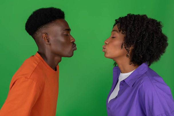 Szczęśliwy młody Afroamerykanin chce pocałować kobietę w luźnym, odizolowanym na zielono tle studia, zbliżenie, profil. Randka razem, związek, miłość i romantyczna rodzina - Zdjęcie, obraz