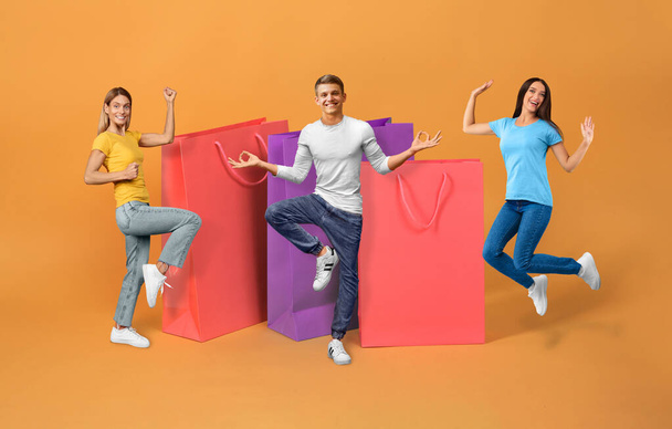 Great Discount. Happy european friends trio celebrating shopping and sale season, jumping and posing near large paper shopper bags over orange studio background. Black friday offer ad. Collage - Fotoğraf, Görsel