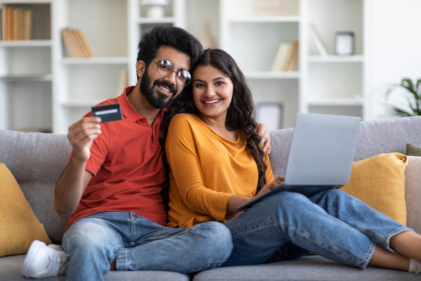Koncepcja handlu elektronicznego. Uśmiechnięta młoda Hinduska małżonka korzystająca z laptopa i karty kredytowej w domu, Szczęśliwa Millennial Eastern para polecająca płatności online, Siedząca na kanapie w salonie, Wolna przestrzeń - Zdjęcie, obraz