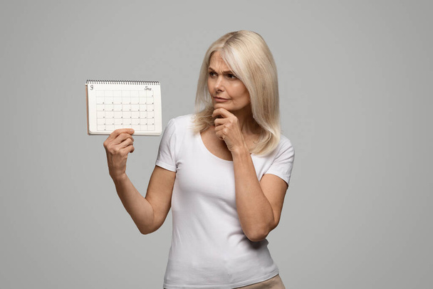 Menopauza koncepció. Töprengő érett nő néz üres menstruációs naptár, feldúlt Senior Nő szenved hiánya menstruáció, miután klimatikus tünetek, pózol szürke háttér, másolás tér - Fotó, kép