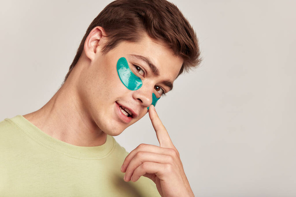 A millennial guy with brown hair practicing skincare with under eye hydrogel patches in a studio. Young smiling caucasian man applying cosmetic patches on his face. Mens self-care - Photo, Image