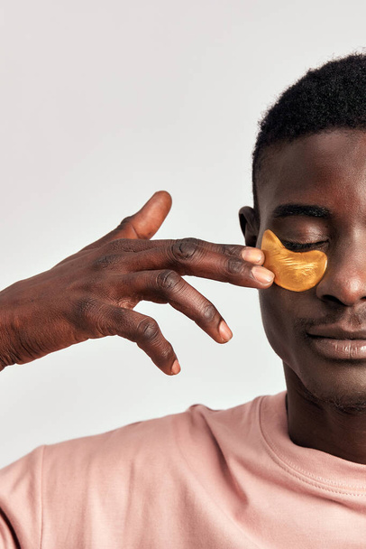 Africký tisíciletý chlap praktikující péči o pleť a péči s pod očima hydrogelové náplasti ve studiu. Mladý usmívající se černoch nanáší kosmetické náplasti na obličej, aby si udržel zdravý vzhled - Fotografie, Obrázek