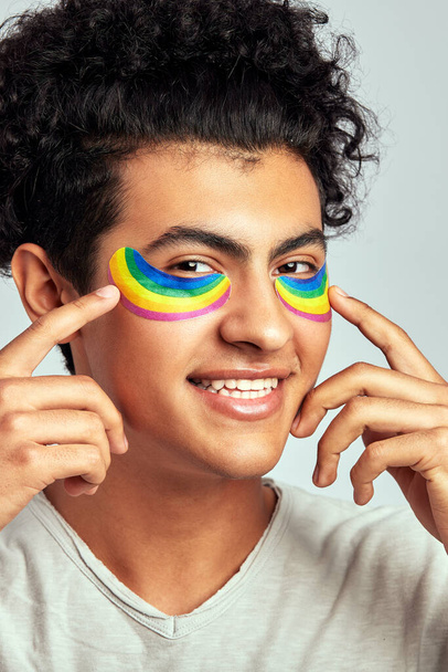 A young handsome guy with black curly hair applying cosmetic patches of rainbow color under his eyes in studio. Swarthy smiling man practicing skincare to maintain his youthful and healthy appearance - Fotografie, Obrázek