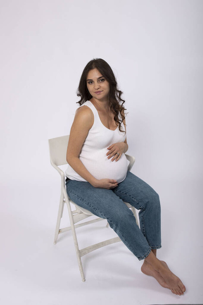 妊娠したブルネット 長い髪の女性. 期待する母親は,椅子に座っている間,優しく彼女の腫れに触れます. スタジオライト,ホワイトバック。 赤ちゃんを待っているコンセプト,健康な妊娠,9ヶ月 . - 写真・画像