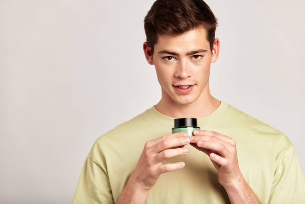Young guy holding a vial of facial mask cream in his hands before applying it on his face during his skincare routine. Man caring for the skin to maintain his youthful and healthy appearance - Photo, Image