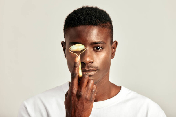 Czarny facet z młodzieńczą skórą, używający jadeitowego wałka do twarzy, by utrzymać skórę miękką i gładką. Przystojny młody Afrykanin utrzymujący swoje naturalne piękno z rutyną pielęgnacji skóry. Samowystarczalność mężczyzn - Zdjęcie, obraz