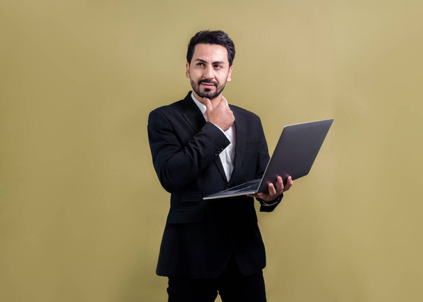 Erfolgreicher Geschäftsmann im schwarzen Anzug mit innovativem Tech-Konzept, stehend in Pose, Laptop in der Hand und aufgeregt lächelnd auf Copyspace-Hintergrund für Promotion oder Werbung. Inbrünstig - Foto, Bild