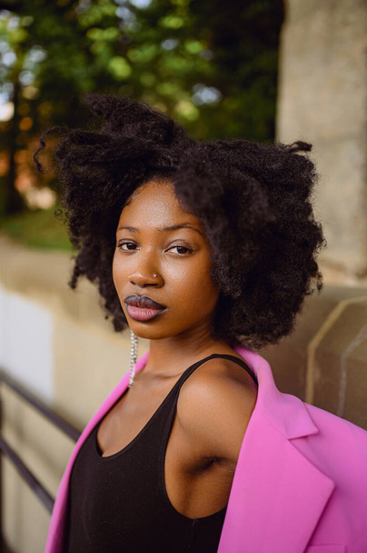 Retrato de estilo urbano al aire libre de moda de la hermosa joven afroamericana posando afuera en el paisaje urbano de la ciudad el día de verano con chaqueta rosa. Atractiva hembra negra, concepto de diversidad - Foto, imagen