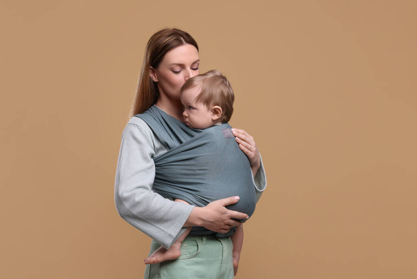 Mother holding her child in baby wrap on light brown background - Фото, изображение