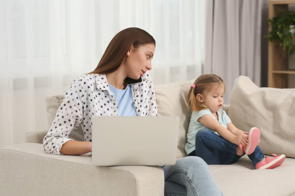 Woman working remotely at home. Busy mother watching over her daughter on sofa in living room - Foto, Bild