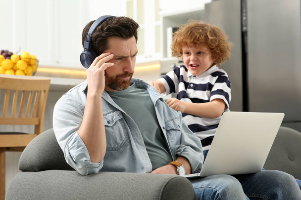 Little boy bothering his father at home. Man with laptop and headphones working remotely - Foto, Bild