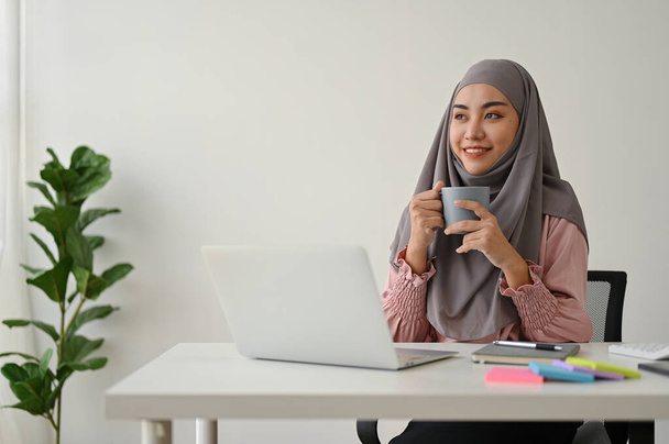 Νεαρή γοητευτική Μουσουλμάνα επιχειρηματίας που πίνει καφέ ενώ δουλεύει με φορητό υπολογιστή στο γραφείο.  - Φωτογραφία, εικόνα