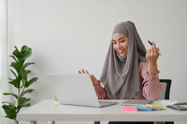 若いアジアのイスラム教徒のビジネスマンは、彼女のチームとビデオ会議を持っています,オフィスルームに座っています.  - 写真・画像