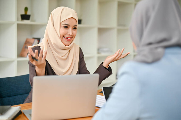 Itsevarma ja ammattimainen aasialainen muslimiliikenainen jakaa ideoitaan ja keskustelee työstään kollegansa kanssa toimistossa.. - Valokuva, kuva