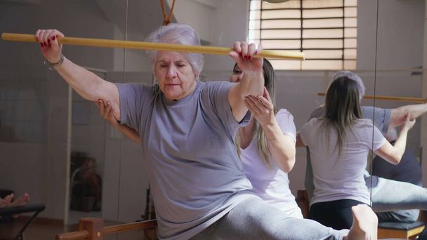 Εκπαιδευτής πιλάτες καθοδηγεί μια ηλικιωμένη γυναίκα στην τάξη φυσιοθεραπείας με ξύλινο ραβδί και τεντωμένο πόδι - Φωτογραφία, εικόνα