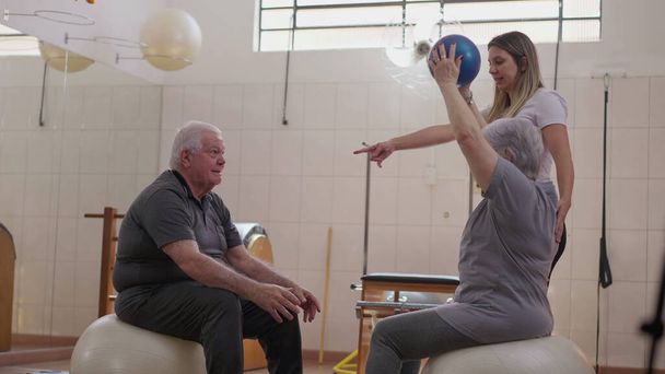 Пожилая пара, тренирующаяся вместе с помощью тренера по пилатесу во время сеанса физиотерапевта - Фото, изображение