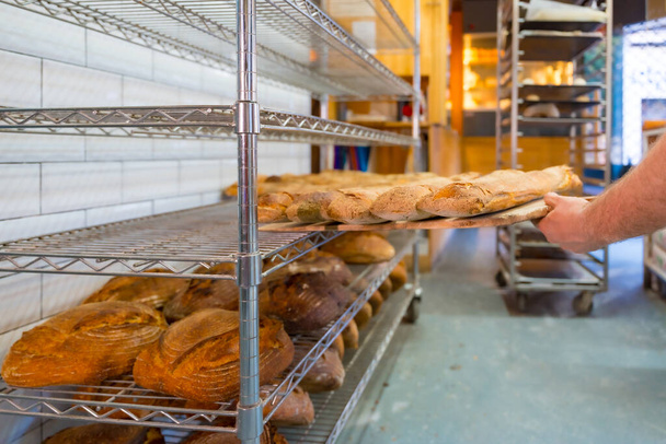 Baker of bakery in the artisan workshop workshop placing the loaves of the oven on the shelves - Zdjęcie, obraz