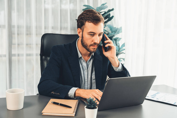 Hardwerkende en ijverige zakenman werkt aan zijn laptop terwijl hij praat met klanten op zijn mobiele telefoon, presentatie van moderne multitasking kantoormedewerker levensstijl. Entiteit - Foto, afbeelding