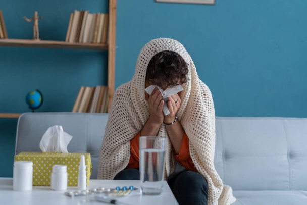 Chory młody arabczyk siedzący na kanapie i kichający bibułą w domu. Mężczyzna dmucha w nos, kaszle lub kicha w domu, cierpi na grypę. Pojęcie zimna i gorączki - Zdjęcie, obraz