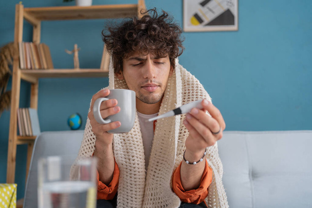 Nemocný muž z Blízkého východu přikrytý kostkovaným sedí na pohovce, pije horký čaj a drží teploměr, mladý nemocný arabský muž má vysokou tělesnou teplotu, příznaky chřipky. Kopírovat prostor - Fotografie, Obrázek