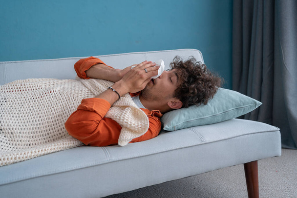 Malo joven árabe acostado en el sofá en casa bajo una manta a cuadros, estornudando en una servilleta, se siente mal, necesita tratamiento y medicamentos. Concepto de estornudo, secreción nasal y tos. Copiar espacio - Foto, imagen