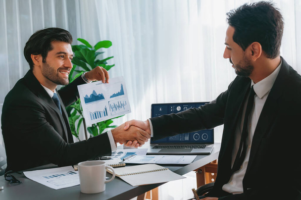 Closeup professional businessman shaking hands over desk in modern office after successfully analyzing pile of dashboard data paper as teamwork and integrity handshake in workplace concept. fervent - Photo, Image