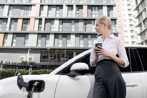 Businesswoman drinking coffee, leaning on electric vehicle recharging at public charging station with residential apartment condos building in background as progressive lifestyle by eco-friendly car. - Foto, immagini