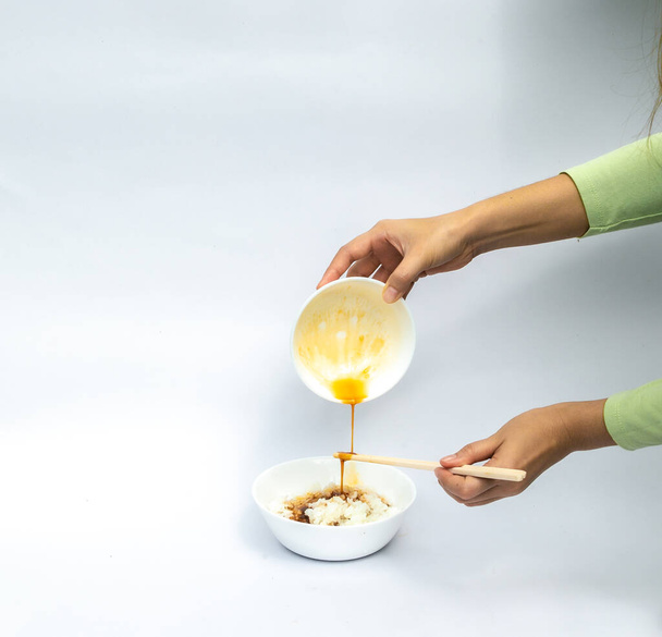 Las manos de las mujeres asiáticas sostienen el huevo fresco se mezcla con salsa de soja y sostienen el palillo mientras se vierte en el tazón de arroz de Japón sobre fondo blanco - Foto, Imagen