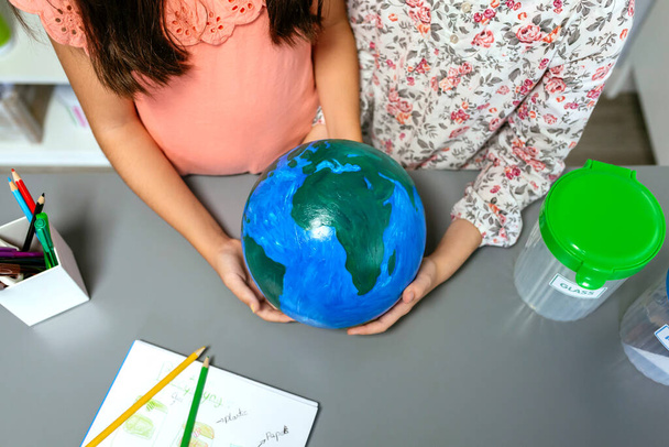 Bovenaanzicht van twee onherkenbare vrouwelijke studenten die een handgemaakte wereldbol in hun handen houden in het ecologie klaslokaal. Duurzaam leefstijlonderwijs. - Foto, afbeelding
