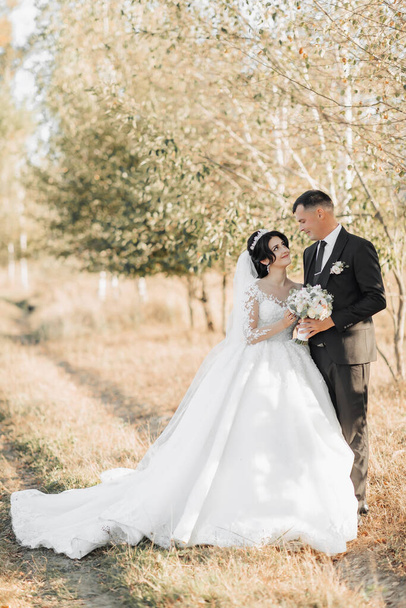 Wedding photo in nature. The groom in a black suit and the bride in a white dress with a train are standing in a field against the background of trees, the bride and groom are looking at each other and holding a bouquet. Portrait. Bright summer weddi - Photo, image