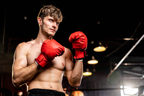 Boxing fighter shirtless posing, caucasian man boxer wearing red glove in defensive guard stance ready to fight and punch at gym with kick bag and boxing equipment in background. Impetus - Photo, Image