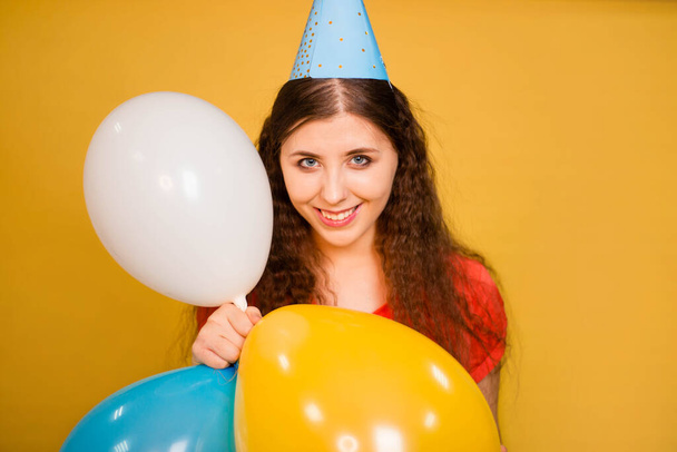 Portrait of a young woman in a festive cap with multi-colored balloons in her hand isolated on a yellow background. Noisy birthday concept. - Photo, image