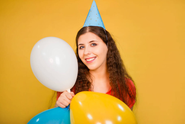Retrato de uma jovem mulher em um boné festivo com balões multicoloridos em sua mão isolados em um fundo amarelo. Conceito de aniversário barulhento. - Foto, Imagem