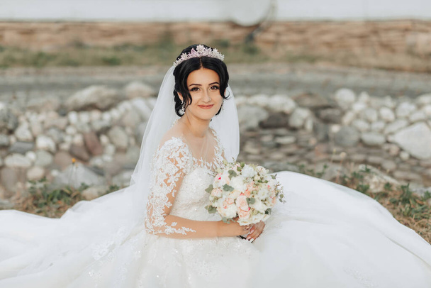 Porträt der Braut in der Natur. Eine brünette Braut mit Krone auf dem Kopf sitzt in einem weißen, voluminösen Kleid, posiert und hält einen Strauß in den Händen. Schönes Haar und Make-up. Hochzeitsshooting - Foto, Bild