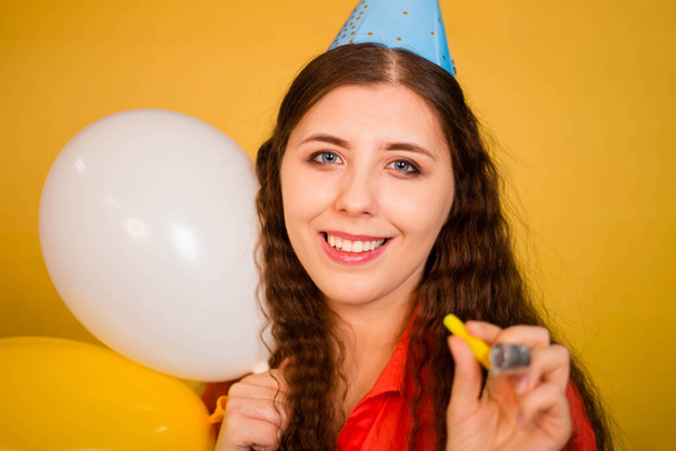 Portrait of a young woman in a festive cap with multi-colored balloons in her hand isolated on a yellow background. Noisy birthday concept. - Zdjęcie, obraz